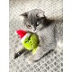 Graue Katze liegt auf einem gemusterten Teppich und hält ein grünes Grinch-Plüschtier mit rotem Weihnachtsmannhut.