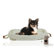 Ein schwarzer und brauner Chihuahua ruht auf einem gemütlichen, gestrickten Haustierbett mit einem hellen Kissen, begleitet von einem kleinen beigen Spielzeug mit einem Herz-Ausschnitt.