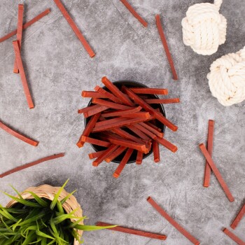 Eine schwarze Schüssel, gefüllt mit dünnen, roten Haustierleckereien, steht in der Mitte einer grauen Oberfläche, umgeben von verstreuten Leckerli-Sticks und zwei weißen Seilspielzeugen.