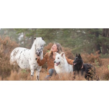 Eine Frau in einem braunen Hemd sitzt in einer natürlichen Umgebung mit einem weiß gescheckten Pony, einem weißen Hund und einem schwarzen Hund, die alle glücklich aussehen.