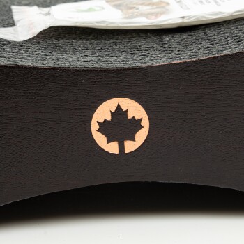 Nahaufnahme eines dunklen Holz-Haustierprodukts mit einem geschnitzten Ahornblatt-Logo, mit einem teilweise sichtbaren Paket von Haustierleckerlis im Hintergrund.