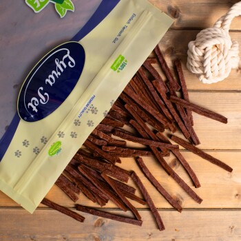 Eine Tüte mit Haustierleckerlis, die braune Jerky-Streifen auf eine Holzoberfläche fallen lässt, mit der Aufschrift "Premium Qualität" und "100% natürlich," mit einem Seilspielzeug in der Nähe.