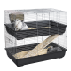 Two-level wire rabbit cage with black plastic trays, ramp, hay and two rabbits (white-gray upper, dark-gray lower).