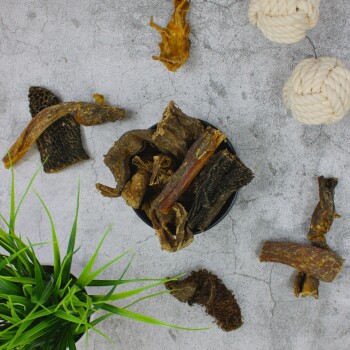 Eine schwarze Schüssel gefüllt mit verschiedenen getrockneten Haustierleckerlis auf einer grauen Oberfläche, mit grünem Gras und zwei weißen Seilspielzeugen in der Nähe.