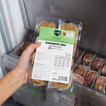 Hand hält transparente Schale mit gefrorenem Hundefutter, beschriftet 'Komplett‑Mix Huhn & Lachs' (400 g), in einem Gefrierschrank