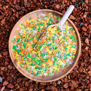 Bunte Haustierbettwäsche aus kleinen, zerrissenen Flocken in einer Holzschüssel mit einer Schaufel, vor einem Hintergrund aus rötlichem Kies.