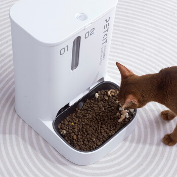 Braune Katze frisst Trockenfutter aus einem weißen automatischen Futterspender mit zwei Einläufen, beschriftet mit '01' und '02'; der Schriftzug 'PETKIT' ist sichtbar.