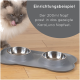 Eine flauschige Katze leckt sich die Lippen neben einer grauen Futtermatte mit zwei Edelstahlschalen, beschriftet mit "KaraLuna Napfset."