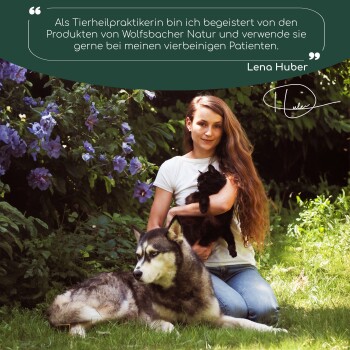 Eine Frau im Freien mit Blumen hält eine schwarze Katze, während ein wolfähnlicher Hund neben ihr liegt. Text darüber bewirbt Wolfsbacher Naturprodukte.