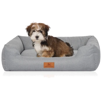 Kleiner braun-weißer, flauschiger Welpe sitzt in grauem Hundebett mit erhöhten Seitenpolstern und Lederetikett 'KNUFFELWUFF'.
