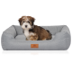 Kleiner braun-weißer, flauschiger Welpe sitzt in grauem Hundebett mit erhöhten Seitenpolstern und Lederetikett 'KNUFFELWUFF'.