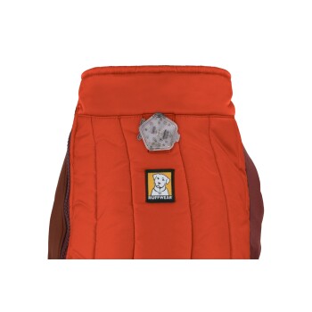 Eine Nahaufnahme einer leuchtend orangefarbenen Hundejacke mit einem Ruffwear-Logo-Patch und einem reflektierenden Lichtanhänger am Kragen.
