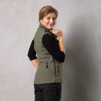 Eine Frau in einer grünen Weste und einem schwarzen Langarmshirt posiert mit dem Rücken zur Kamera und zeigt die Reißverschlüsse der Weste.