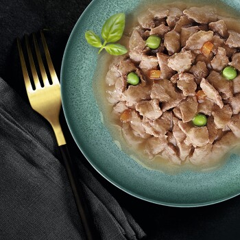 Mokra karma dla kotów w zielonej misce, z kawałkami mięsa i zielonymi groszkami, udekorowana świeżymi liśćmi bazylii, z złotym widelcem obok.