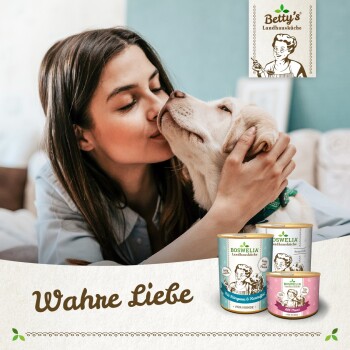Eine Frau interagiert mit einem Golden Retriever Welpen in der Nähe von drei Dosen von Bettys "Boswellia Landhausküche" Haustierfutter, beschriftet mit "Wahre Liebe."