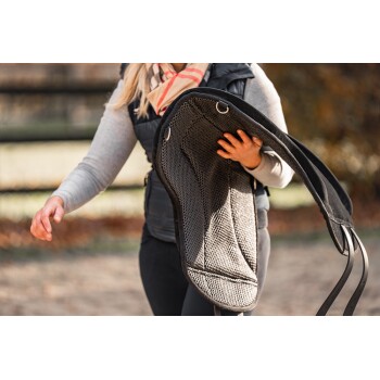 Eine Frau in einem grauen Pullover und einer schwarzen Weste trägt ein schwarzes Pferdesattelpad mit Riemen, vor einem verschwommenen Hintergrund im Freien.