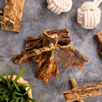 Natürliche Haustierkausnacks und Spielzeuge auf einer grauen Oberfläche, darunter getrocknete Rindenleckerlis, weiße Seilspielzeuge und eine kleine grüne Pflanze in einem Korb.