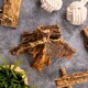 Natürliche Haustierkausnacks und Spielzeuge auf einer grauen Oberfläche, darunter getrocknete Rindenleckerlis, weiße Seilspielzeuge und eine kleine grüne Pflanze in einem Korb.