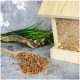 Ein hölzerner Vogelhausfütterer, gefüllt mit Samen, neben einer Schaufel, die Samen auf einer strukturierten Oberfläche verschüttet, mit Gras und Treibholz im Hintergrund.