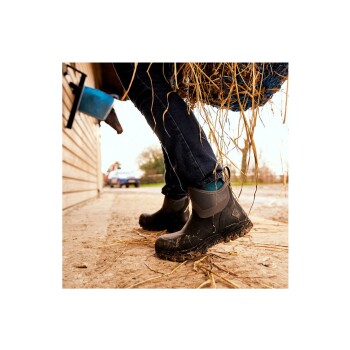 Eine Nahaufnahme von schlammigen schwarzen Gummistiefeln, die von einer Person getragen werden, die ein Bündel Heu trägt, mit einer rustikalen Scheune und einem blauen Truck im Hintergrund.