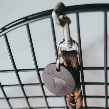 Eine Nahaufnahme eines braunen Leder-Hundemarkenanhängers, der an einer geflochtenen Leine hängt, die von einem schwarzen Metallgitter hängt.