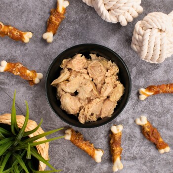 Eine schwarze Schüssel, gefüllt mit zerkleinertem, feuchtem Haustierfutter, steht auf einer grauen Oberfläche, umgeben von Hundekauspielzeugen und einem Seilspielzeug, mit einer kleinen Pflanze in der Nähe.