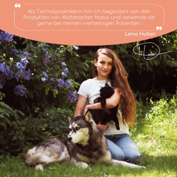 Eine Frau hält eine schwarze Katze neben einem großen Hund in einem Garten mit lila Blumen. Der Text darüber lautet: "Als Tierheilerin liebe ich die Produkte von Wolfsbacher Natur."