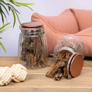 Zwei Glasbehälter gefüllt mit Hundeleckerlis, einer umgekippt und verschüttet Leckerlis, neben einem Seilspielzeug auf einem Holztisch und einem rosa Haustierbett im Hintergrund.