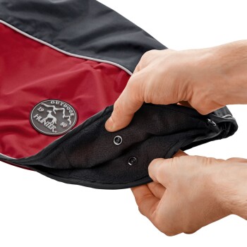 Eine Hand hält einen schwarz-roten Hundemantel und zeigt das Innenfutter, Druckknöpfe und das Logo "OUTDOOR HUNTER."
