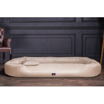 Ein plüschiges beiges Haustierbett mit einem kleinen Kissen, vor einer dunklen Paneelwand und einem Holzboden, ideal für den Komfort von Haustieren.