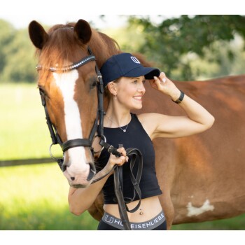 Eine Frau in einem schwarzen Crop-Top und einer Kappe steht neben einem braunen Pferd, hält die Zügel und passt ihren Hut an, mit einem grünen Feld im Hintergrund.