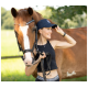 Eine Frau in einem schwarzen Crop-Top und einer Kappe steht neben einem braunen Pferd, hält die Zügel und passt ihren Hut an, mit einem grünen Feld im Hintergrund.