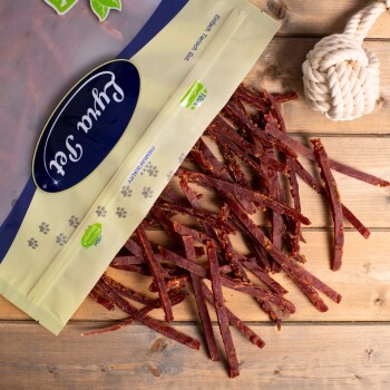 Eine Tüte mit Haustierleckereien mit der Aufschrift "Premium Qualität" mit einem Logo, die Streifen von Trockenfleisch auf eine Holzoberfläche fallen lässt, neben einem Seilspielzeug.