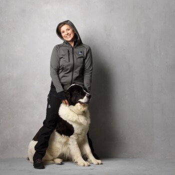 Eine Person in einem grauen Reißverschluss-Hoodie und schwarzen Hosen sitzt hinter einem großen, flauschigen schwarz-weißen Hund, beide vor einem grauen Hintergrund.