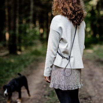 Eine Frau mit lockigem Haar trägt einen hellen Pullover und einen Blumenrock und führt einen Hund auf einem Waldweg, mit einer Leine, die an ihrer Kleidung befestigt ist.