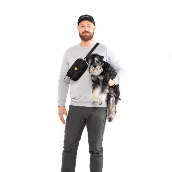 Eine Person in einem grauen Sweatshirt und schwarzer Mütze hält einen kleinen schwarz-grauen Hund in einem Arm, während sie eine schwarze Umhängetasche trägt.