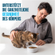 Mann streichelt eine getigerte Katze, die aus einer weißen Trockenfutterschüssel frisst; sichtbarer Text: UNTERSTÜTZT DIE GANZHEITLICHE GESUNDHEIT DES KÖRPERS