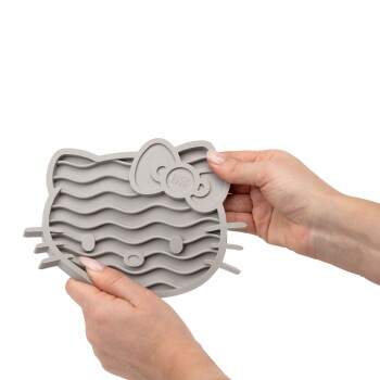 Eine Person hält eine graue Silikon-Haustierfütterungsmatte in Form eines Hello Kitty-Gesichts, mit wellenförmigen Linien und einer Schleife, beschriftet mit "LUCY BALU."