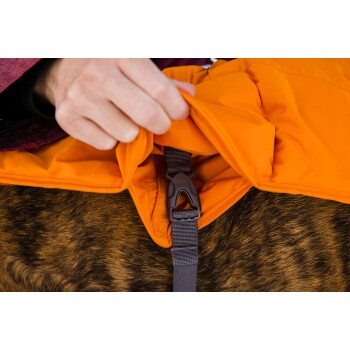 Eine Nahaufnahme einer Hand, die eine orangefarbene Haustierjacke mit einer braunen Schnalle befestigt, die auf dem Fell eines Hundes ruht.