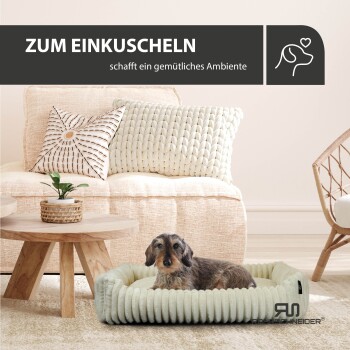 Ein kleiner Hund entspannt sich auf einem runden Bett in einem gemütlichen Wohnzimmer mit einem hellen Sofa, Kissen und einer Pflanze. Text lautet: "ZUM EINKUSCHELN."