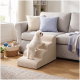 Kleiner weißer Hund steigt die beige Haustiertreppe zu einem hellgrauen Sofa mit pastellfarbenen Kissen, Spielzeugkorb und Decke in der Nähe hinauf.
