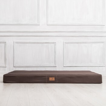 Braunes Haustierbett mit einem Lederetikett, auf einem hellen Holzboden gegen eine weiße Wand platziert.