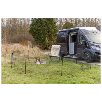 Ein Hund ruht auf einer Decke innerhalb eines tragbaren Haustierzauns, mit einem schwarzen Wohnmobil, das in der Nähe auf einem Grasbereich geparkt ist.