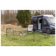 Ein Hund ruht auf einer Decke innerhalb eines tragbaren Haustierzauns, mit einem schwarzen Wohnmobil, das in der Nähe auf einem Grasbereich geparkt ist.