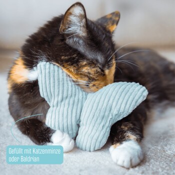 Eine dreifarbige Katze kuschelt mit einem hellblauen, schmetterlingsförmigen Spielzeug, beschriftet mit "Gefüllt mit Katzenminze oder Baldrian", auf einer weichen Oberfläche.