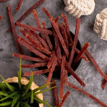 Eine schwarze Schüssel gefüllt mit dünnen, zähen Streifen von Haustier-Jerky, umgeben von einer kleinen grünen Pflanze und zwei geknoteten Seilspielzeugen auf einer grauen Oberfläche.