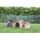 Ein brauner und grauer Hase sitzt neben einem Holzhasenhaus mit einem schrägen Dach und einem kleinen Fenster, umgeben von grünem Gras und einem Drahtzaun.