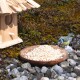 Ein Blaumeise pickt an einem Haufen Vogelfutter auf einem Holzbrett, neben einem Holzvogelhaus, umgeben von Kieselsteinen und Moos.