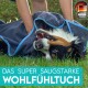 Eine Person wickelt spielerisch einen glücklichen Hund in ein weiches, dunkles Handtuch mit blauen Akzenten auf grünem Gras. Der Text lautet "DAS SUPER SAUGSTARK WOHLFÜHLTUCH."