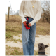 Eine Person in einem cremefarbenen Pullover und blauen Jeans hält eine rote Leckerlitasche für Haustiere, während sie im Freien neben einem grauen Hund steht, der nach oben schaut.